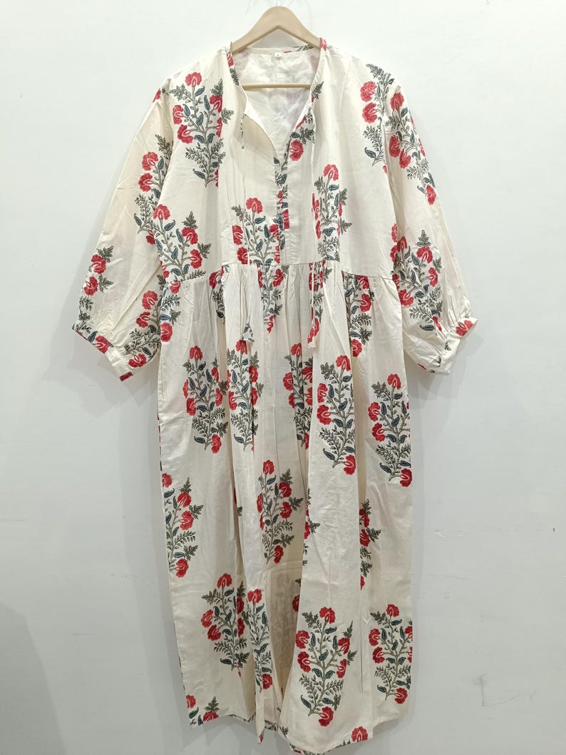 May include: A white cotton dress with a floral print in shades of red and green. The dress has a V-neckline and a tie at the neckline. The sleeves are three-quarter length and the dress has a gathered skirt.