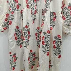 May include: A white cotton dress with a floral print featuring red and green flowers. The dress has a gathered waist and long sleeves.