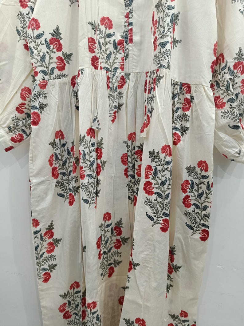 May include: A white cotton dress with a floral print featuring red and green flowers. The dress has a gathered waist and long sleeves.