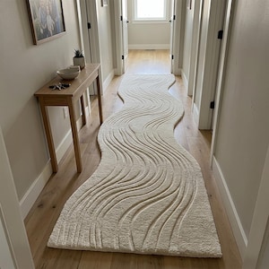 Peut inclure: Un long tapis de couloir couleur crème avec un motif sculpté, ondulé et texturé qui imite l'eau courante. Le tapis présente une forme organique distincte, non rectangulaire, qui serpente sur le sol d'un couloir en bois clair.