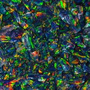 May include: A close-up view of a collection of iridescent black opal gemstones. The stones are irregularly shaped and range in size from 3 to 6 mm. The opals display a vibrant play of color, with flashes of green, blue, yellow, and red.