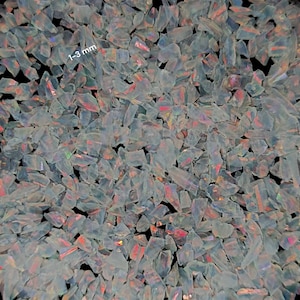 May include: A collection of small, translucent opal chips with an iridescent play of colour, featuring flashes of red, orange, and blue. The individual fragments are irregular in shape and size, with the text 1-3 mm visible on the surface.