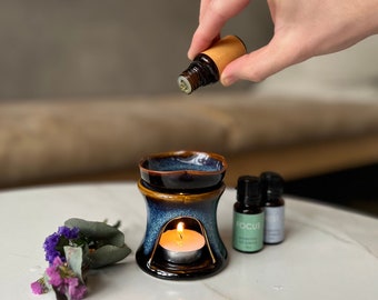 May include: A blue and brown ceramic essential oil diffuser with a lit candle inside. A hand is pouring oil from a small bottle. Two additional essential oil bottles and a small floral arrangement are also present.