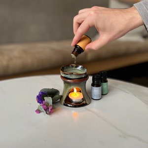 May include: A ceramic oil diffuser with a lit candle inside. Three small bottles of essential oil are on the table, labelled "REST", "FOCUS", and "MINDFULNESS".