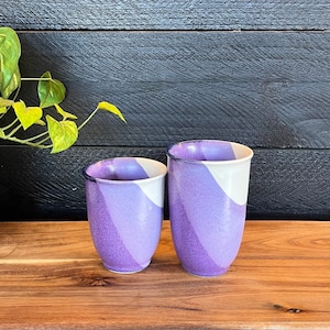 May include: Two purple and white ceramic mugs with a wavy design. The mugs are sitting on a wooden surface.