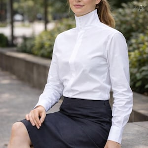 Puede incluir: Una camisa blanca de manga larga con cuello alto y detalles de botones. La camisa se combina con una falda gris oscuro. La imagen presenta un estilo clásico y elegante, adecuado para entornos profesionales o formales.