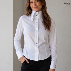 Puede incluir: Camisa blanca de manga larga con cuello alto y botones plateados. La camisa tiene un corte entallado y puños abotonados. El cuello presenta tachuelas plateadas decorativas. Se combina con pantalones negros.