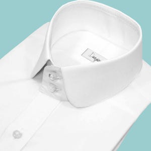 May include: A crisp white dress shirt with a rounded collar and two small buttons. The shirt has a classic design and the label reads "Darje". The shirt is made of a smooth, likely cotton, fabric.