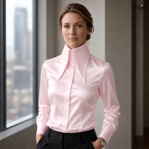 May include: A light pink satin button-down shirt with a high, pointed collar. The long-sleeved shirt is paired with black pants. The image is taken indoors, with a window in the background.