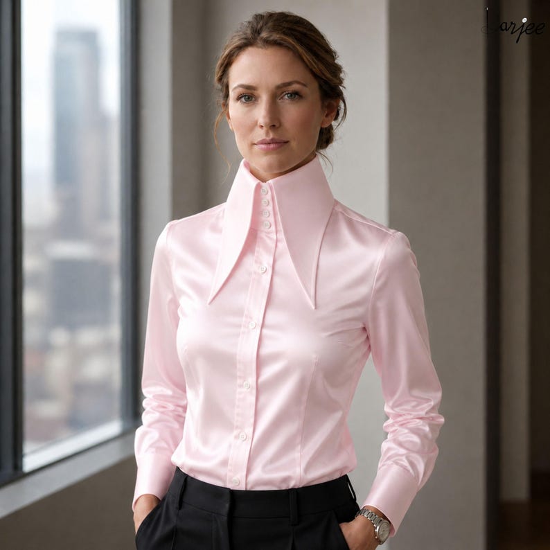 May include: A light pink satin button-down shirt with a high, pointed collar. The long-sleeved shirt is paired with black pants. The image is taken indoors, with a window in the background.