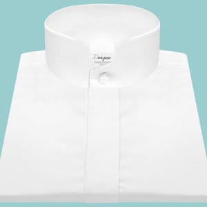 May include: A crisp white shirt with a mandarin collar. The shirt features a button closure and the brand name "Darjee" is visible on the collar. The shirt is neatly folded, and the background is a light teal color.