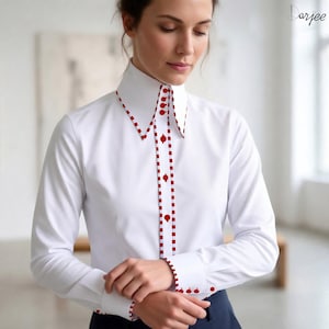 May include: A crisp white long-sleeved button-down shirt featuring a high collar and a central placket adorned with a red and white checkered trim. The shirt includes red buttons along the front and cuffs, with the brand name Darjee visible in the corner.