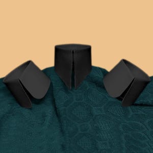 May include: Three black detachable collars, two with rounded corners and one with a pointed front, rest on a textured teal fabric. The collars are made of a smooth, matte material and are set against a light tan background.
