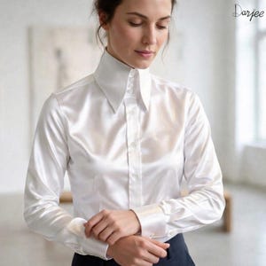 May include: A long-sleeved white satin button-down shirt featuring a structured, pointed collar and a front placket with visible buttons. The fabric has a smooth, reflective finish, and the cuffs are secured with buttons. The text Darjee is visible.