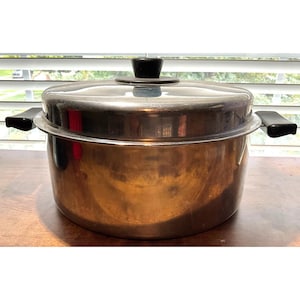 May include: A copper-toned metal steamer pot with a clear glass lid and black handles. The lid has a black knob. The pot sits on a wooden surface in front of a window with blinds.
