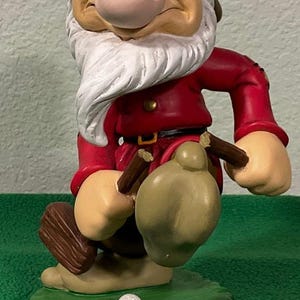 May include: A Grumpy figurine from Snow White wearing a brown hat and red tunic, holding a golf club and ball. The character stands on a green base with a small white golf ball and a hole, featuring the text Walt Disney World printed on the front.