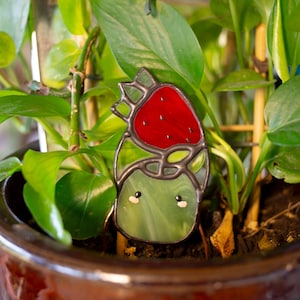 May include: A stained glass figurine of a green creature with a red strawberry on its head. The figurine has white eyes and a black outline. The figurine is sitting in a pot of green plants.
