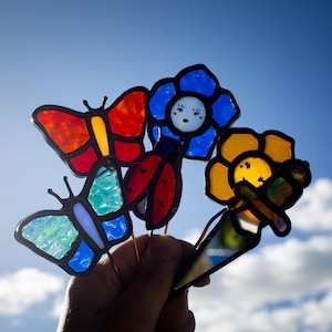 May include: A selection of vibrant stained glass garden ornaments, including butterfly, ladybird, and flower designs. The butterfly stakes have red, orange, and blue wings. The flower stakes are blue and yellow. The stakes are held against a bright blue sky.