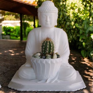Puede incluir: Estatua de Buda de cerámica blanca con una planta de cactus en una maceta blanca con forma de loto. La estatua está sentada sobre una base blanca.