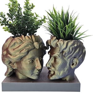 May include: Two ceramic planters shaped like human heads, facing each other. The planters are painted in a muted green and brown color scheme. The planter on the left has a female face and is filled with green leafy plants. The planter on the right has a male face and is filled with green grass.