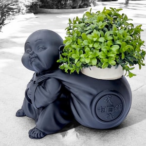 May include: Black ceramic planter shaped like a Buddha carrying a pot with green plants. The pot has a white rim and a Chinese character on the side.