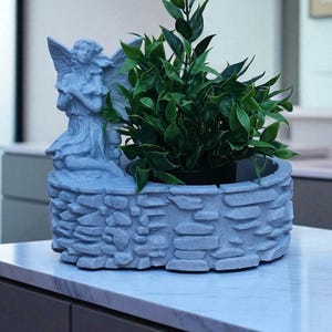 May include: A gray stone planter with an angel statue on the side, filled with green leafy plants. The planter has a textured, stacked stone design. The angel is depicted with wings and a flowing gown. The planter sits on a white countertop.