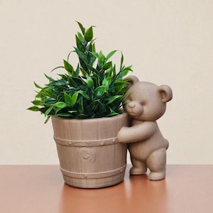 May include: A decorative planter featuring a small, brown teddy bear hugging a wooden-style pot filled with green foliage. The bear is in a standing position, adding a whimsical touch to the design. The pot has a barrel-like appearance.