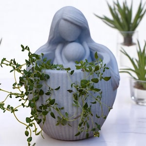 May include: A blue ceramic planter shaped like a woman holding a child, with green plants growing out of the top.