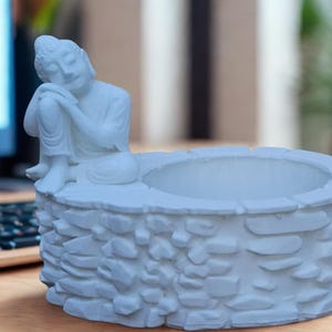 May include: A light blue Buddha statue planter. The planter has a well-like design with a seated Buddha figure resting on the side. The planter is approximately 6 inches wide and 4 inches tall.