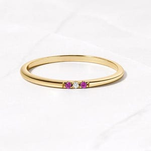 Op de afbeelding: Een delicate gouden ring met drie edelstenen: twee roze robijnen flankeren een enkele witte diamant. De ring is geplaatst op een witte ondergrond, wat het minimalistische ontwerp en de elegante eenvoud benadrukt.