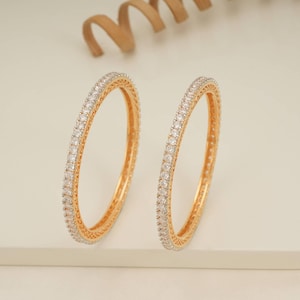 Może przedstawiać: Dwie złote bransoletki z ciągłym rzędem małych, przezroczystych kamieni szlachetnych. Bransoletki mają polerowane złote wykończenie. Kamienie szlachetne błyszczą, odbijając światło. Neutralne tło.