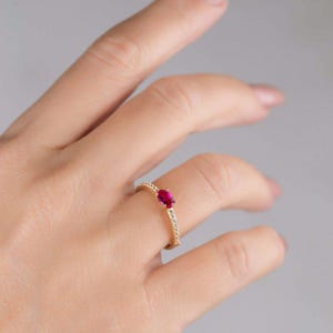 May include: A gold ring featuring a vibrant red oval-cut gemstone as the centerpiece. The band is adorned with small, sparkling clear stones. The ring is displayed on a hand against a neutral background, showcasing its elegant design.