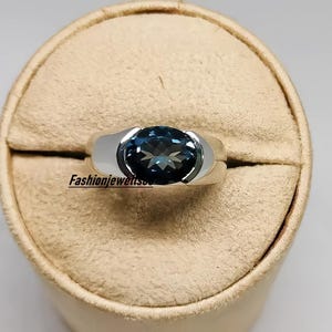 Può includere: Un anello d'argento con una pietra preziosa blu incastonata in una montatura a castone. L'anello è su un espositore beige.