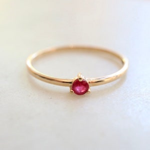 May include: A delicate gold band ring with a single, small, round ruby gemstone.