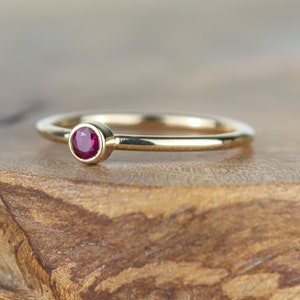 May include: A simple gold band ring with a small, round ruby gemstone set in the center.