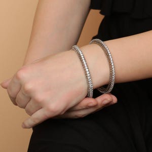 Może przedstawiać: Dwie srebrne bransoletki wysadzane małymi, przezroczystymi kamieniami szlachetnymi. Bransoletki są noszone na nadgarstku, a druga ręka je trzyma. Biżuteria jest umieszczona na czarnym topie i neutralnym tle.