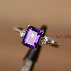 May include: A silver ring featuring a large, rectangular, deep purple gemstone as the focal point. Two smaller, square-shaped clear stones flank the main stone. The ring's band is thin and polished.