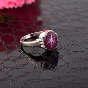 Może przedstawiać: Srebrny pierścionek z dużym, owalnym kamieniem szlachetnym w kształcie rubinu gwiazdowego. Rubin ma sześcioramienny wzór gwiazdy. Pierścionek ma małe, przezroczyste kamienie akcentujące. Pierścionek jest na ciemnym, teksturowanym tle.