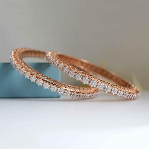 Może przedstawiać: Dwie bransoletki z różowego złota z kwiatowym wzorem. Bransoletki są inkrustowane małymi, błyszczącymi białymi kamieniami szlachetnymi. Wzór przedstawia powtarzający się wzór klastrów w kształcie kwiatów, dodając elegancji biżuterii.