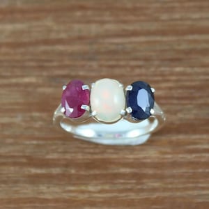 May include: A silver ring featuring three oval-shaped gemstones. The central stone is a white opal, with a red ruby to the left and a blue sapphire to the right.
