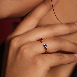 Puede incluir: Un anillo dorado con diseño abierto, con una gema rectangular de rubí y amatista. El anillo se lleva en un dedo, mostrando el diseño y el contraste de color de la joya.