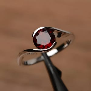 Peut inclure: Une bague en argent avec une pierre précieuse rouge. La bague a un design simple avec une seule pierre sertie dans une bande lisse.