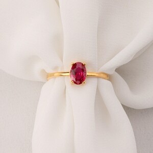 Puede incluir: Un anillo de oro con una gran gema de rubí ovalada. El rubí es de color rojo brillante, engastado en una sencilla banda de oro. El anillo se muestra sobre un fondo de tela blanca. El diseño es elegante.