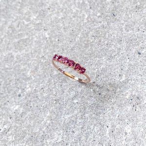 Puede incluir: Un delicado anillo de oro con una fila de piedras preciosas rosas dispuestas en línea recta.