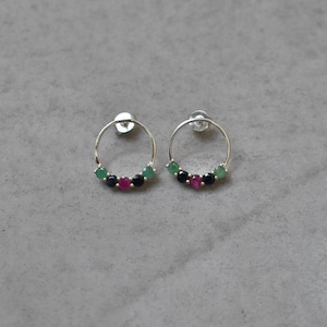 Puede incluir: Un par de pendientes de aro de plata con piedras preciosas verdes, rojas y negras. Los pendientes son pequeños y delicados.
