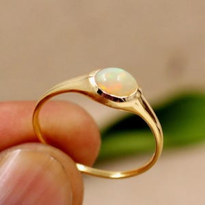 Könnte beinhalten: Ein goldener Ring mit einem glatten, ovalen Opal-Edelstein. Der Opal hat eine milchig-weiße Basis mit orangefarbenen und gelben Lichtreflexen. Das Ringband ist aus einfachem, poliertem Gold.