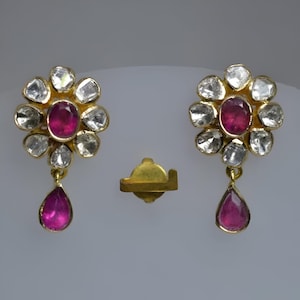 May include: A pair of gold earrings with a floral design. Each earring features a cluster of small, clear stones surrounding a larger, pink stone. A smaller pink stone dangles from the bottom of each earring.