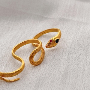 May include: A gold-colored snake ring with a coiled design. The snake's head features a red and a blue gemstone for eyes. The ring is set against a white background.