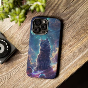 May include: A phone case featuring a dark-colored cat sitting on a planet with a space-themed background. The case has a dark blue border and a multi-lens camera cutout. The artwork includes planets, stars, and nebulas.