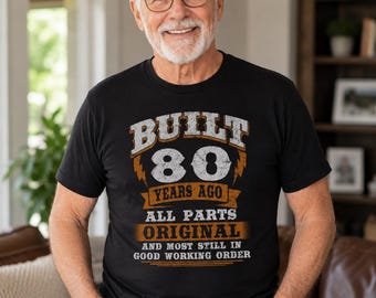 May include: Black t-shirt with the text "BUILT 80 YEARS AGO ALL PARTS ORIGINAL AND MOST STILL IN GOOD WORKING ORDER" in white and orange. The shirt is worn by a person with glasses.